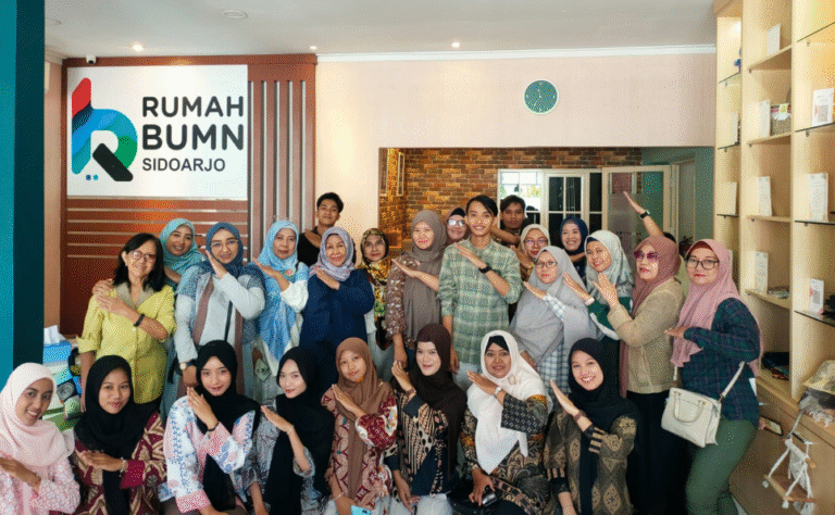 Foto bersama seluruh peserta dan panitia Seminar Power of Visual Content di dalam ruangan Rumah BUMN Sidoarjo dengan latar belakang logo instansi